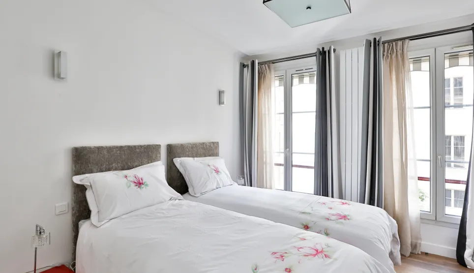 Chambre lumineuse et élégante avec deux lits jumeaux, draps fleuris, têtes de lit capitonnées. Grandes fenêtres et décoration moderne pour un confort de luxe.