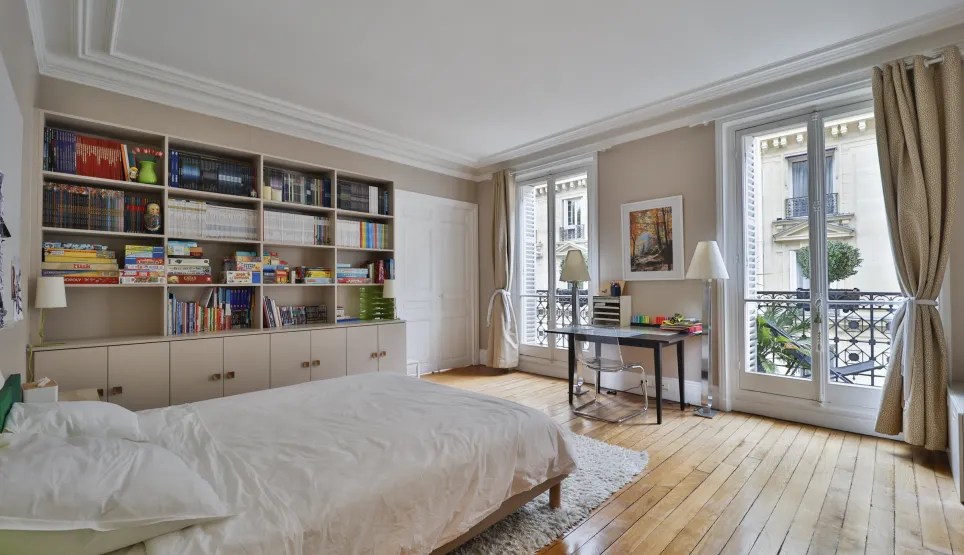 Chambre parisienne lumineuse avec parquet, moulures, bibliothèque intégrée et balcons. Élégance et confort pour un appartement de luxe.