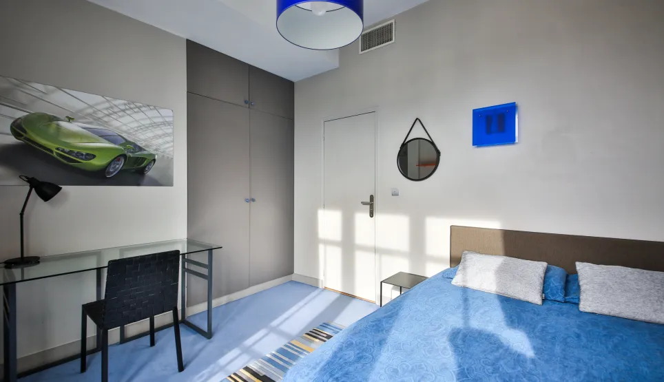 Chambre moderne et lumineuse, design épuré avec lit bleu, bureau verre, placards intégrés et œuvre d'art voiture sport. Ambiance chic.