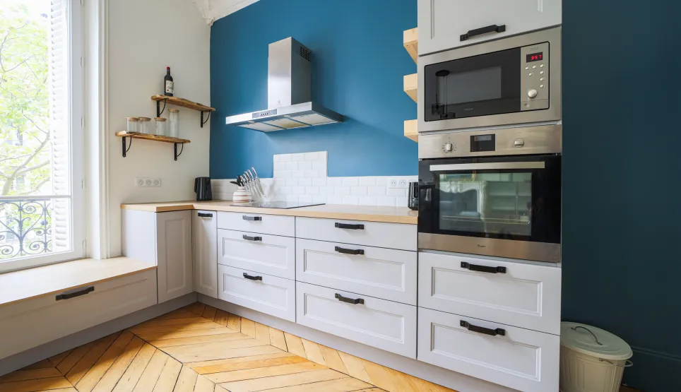 Cuisine moderne et lumineuse, murs bleus, parquet chevrons. Design élégant avec rangements blancs et électroménager intégré. Appartement parisien.