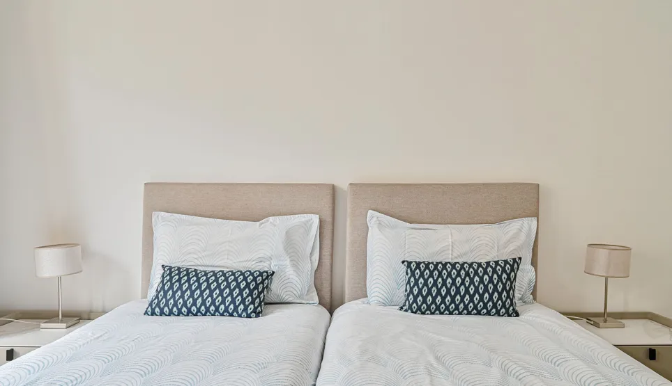 Chambre double luxe avec lits jumeaux, têtes de lit beige, literie épurée et coussins bleus. Design moderne et ambiance sereine pour un séjour raffiné.