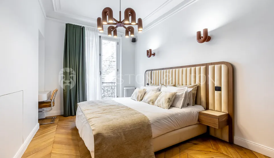Chambre de luxe moderne avec tête de lit capitonnée, parquet chevrons, luminaires design et balcon. Élégance et confort pour un intérieur parisien.
