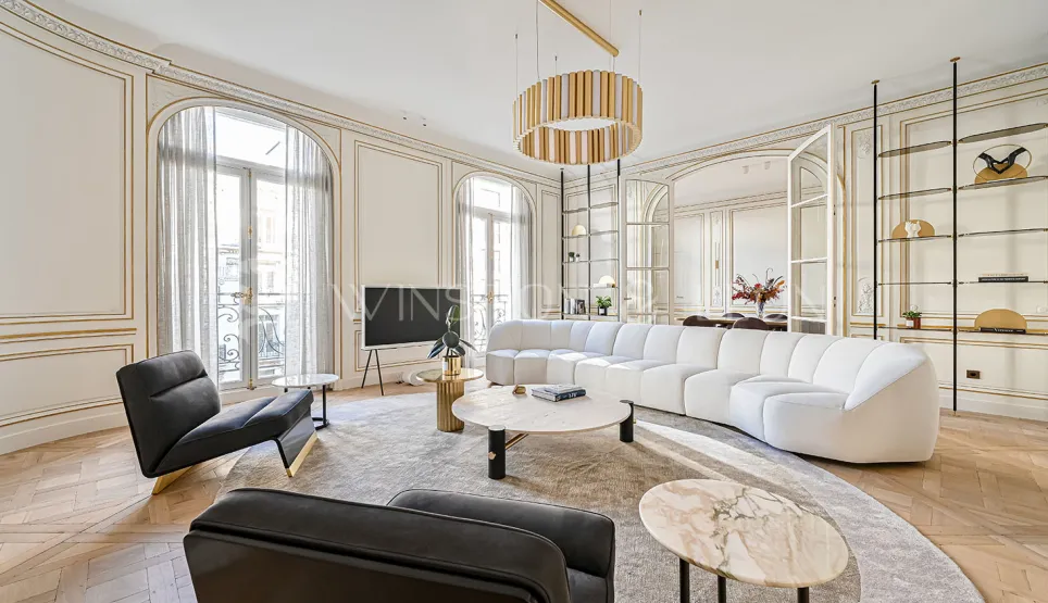 Salon Haussmannien parisien d'exception: moulures dorées, parquet point de Hongrie, canapé design blanc, mobilier luxe et lumière naturelle.