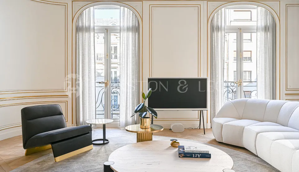 Salon de luxe parisien avec moulures dorées, mobilier design contemporain, grandes fenêtres cintrées et lumière naturelle. Élégance et raffinement.