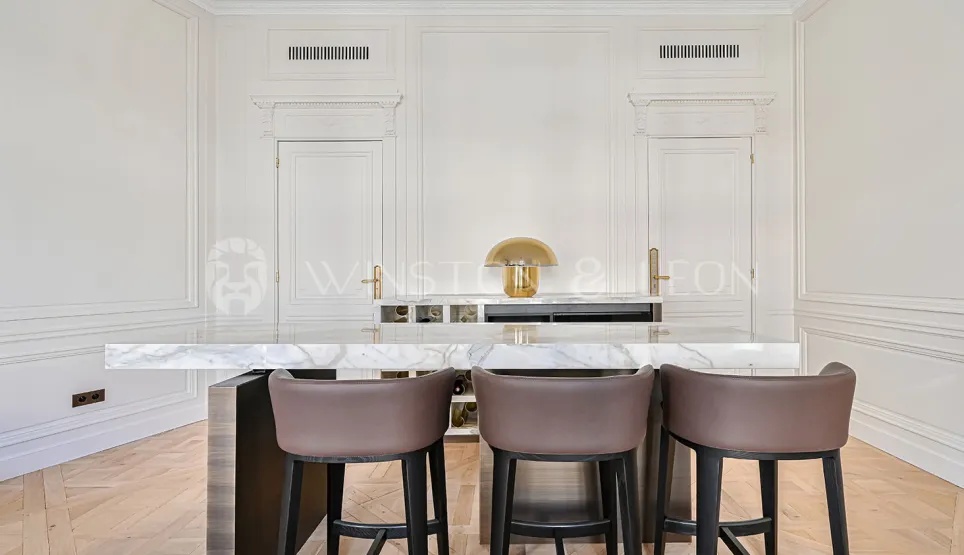 Cuisine de luxe avec îlot marbre, tabourets design. Boiseries élégantes, lampe dorée. Intérieur prestige, appartement parisien chic.