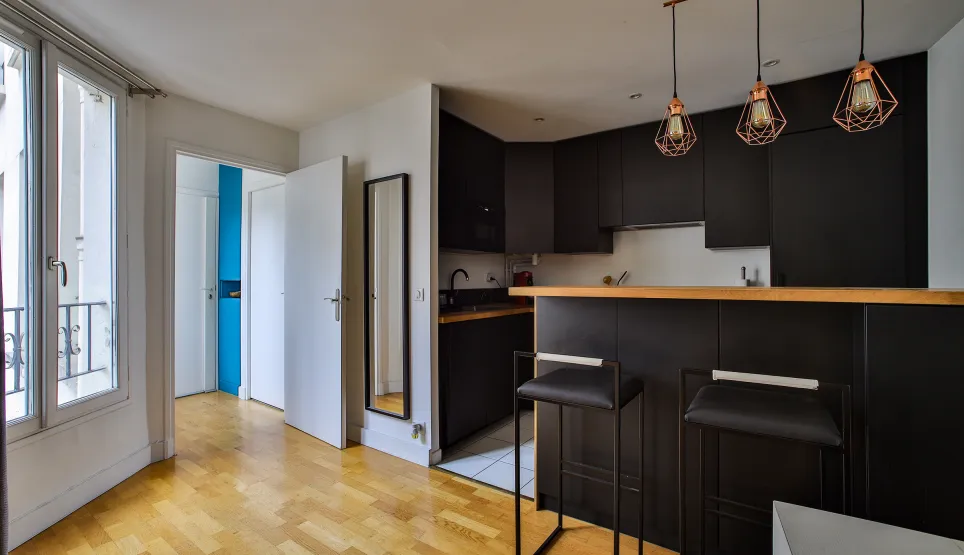 Cuisine moderne noire avec bar, tabourets design, parquet et luminaires cuivrés. Un espace de vie lumineux et élégant.