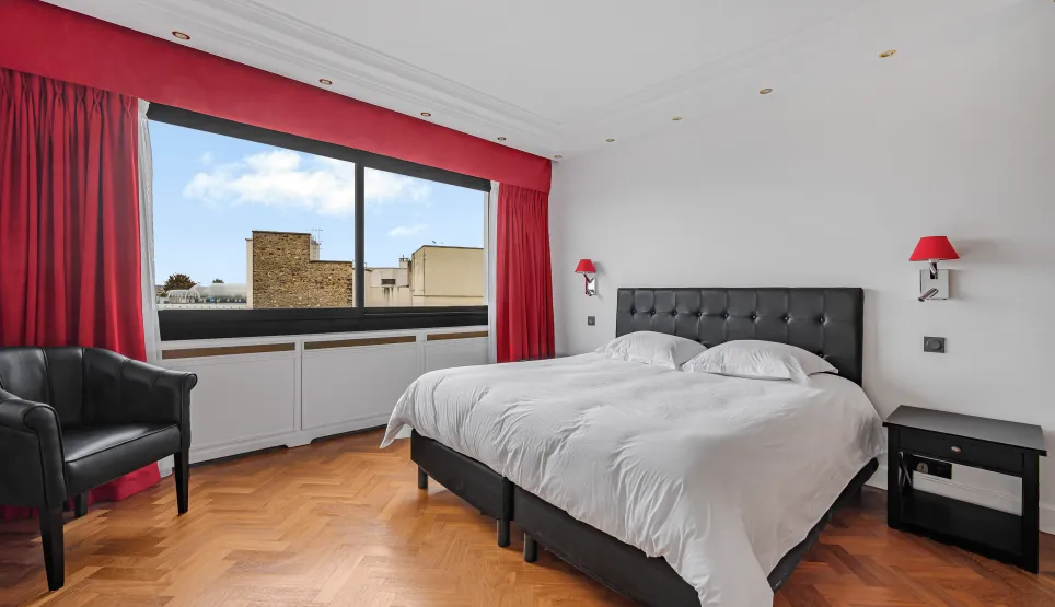Chambre de luxe lumineuse, vue ville, parquet chevrons. Lit double élégant, fauteuil. Parfait pour immobilier haut de gamme.