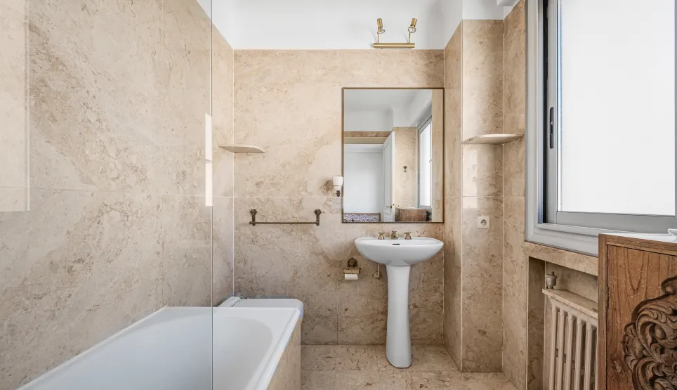 Salle de bain élégante en marbre beige, avec baignoire, lavabo sur colonne et miroir. Design classique et lumineux pour un intérieur de luxe.