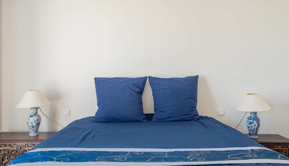 Chambre élégante et confortable avec lit bleu profond, tables de chevet sculptées et lampes en porcelaine. Ambiance raffinée pour propriété de luxe.