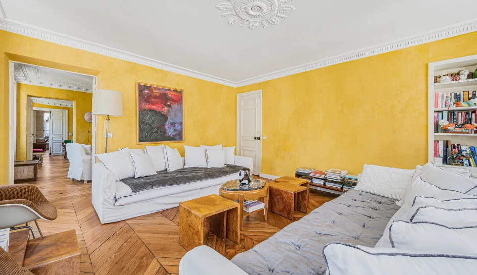Salon de luxe parisien lumineux: murs jaunes, parquet chevrons, moulures, canapés blancs. Élégance et confort.