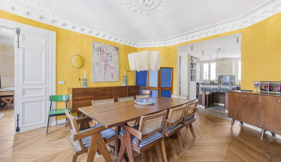 Appartement parisien de luxe: salle à manger lumineuse aux murs jaunes, parquet point de Hongrie, moulures. Cuisine ouverte design. Élégance vibrante.