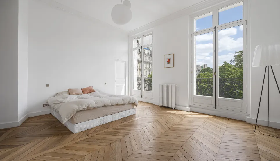 Chambre lumineuse d'appartement Haussmannien. Parquet chevrons, hauts plafonds, grandes fenêtres, vue verdure. Luxe parisien épuré.
