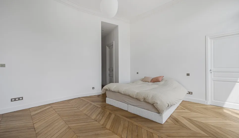 Chambre luxe minimaliste, parquet chevron, murs blancs. Élégance parisienne, luminosité naturelle. Idéal appartement haussmannien.
