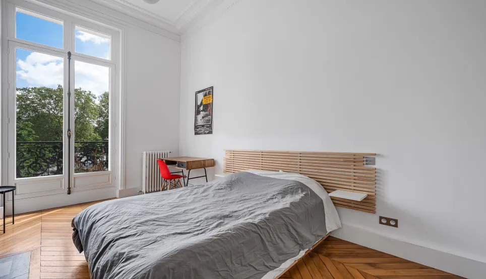 Chambre lumineuse avec balcon vue verdure, parquet chevrons et moulures. Design moderne et élégance parisienne pour un luxe discret.