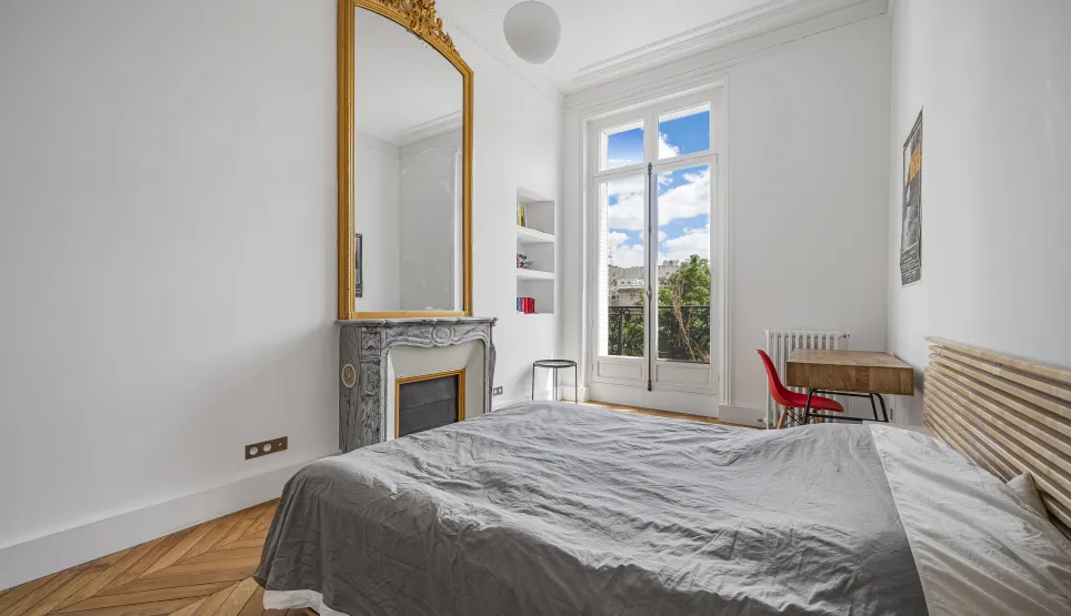 Chambre lumineuse avec parquet chevrons, cheminée marbre, miroir doré et vue dégagée. Charme haussmannien pour appartement de luxe.