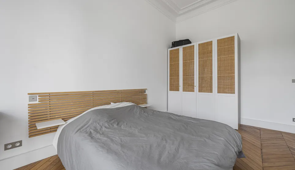 Chambre épurée: tête de lit bois, dressing rotin, parquet chevrons. Ambiance sereine et élégante pour un intérieur de luxe parisien.