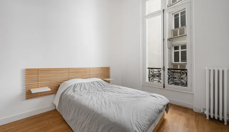 Chambre lumineuse au design épuré, parquet, tête de lit bois moderne, fenêtre parisienne. Appartement de prestige.