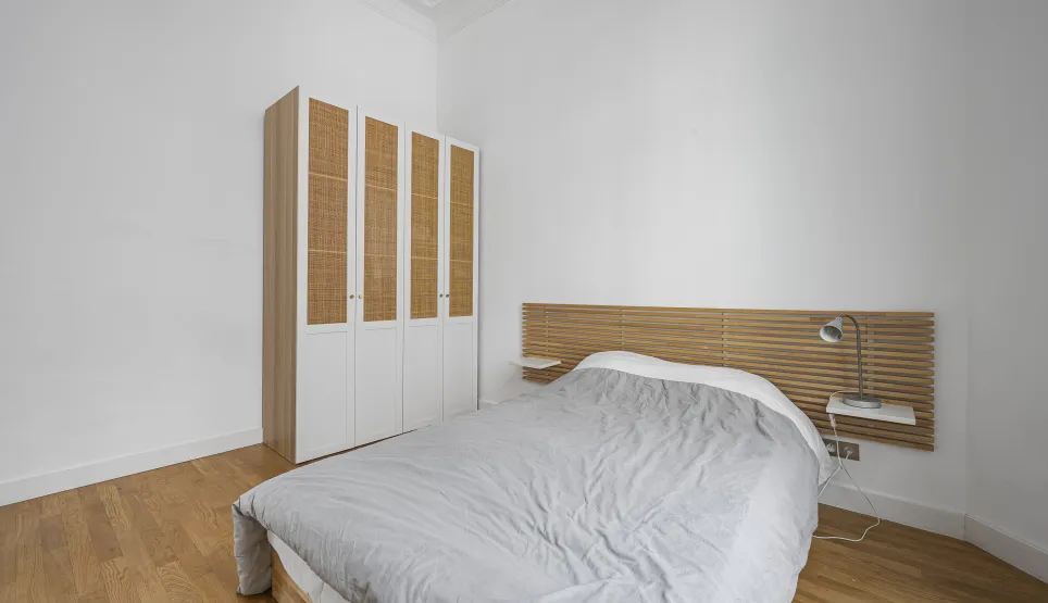 Chambre moderne et lumineuse avec parquet, tête de lit design en bois, et armoire élégante en rotin. Idéal pour appartement de luxe.
