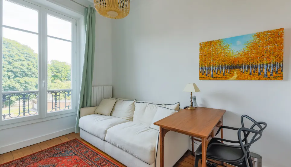 Pièce lumineuse avec vue verdoyante, canapé blanc, bureau bois et art moderne. Ambiance sereine et élégante pour travail ou détente.