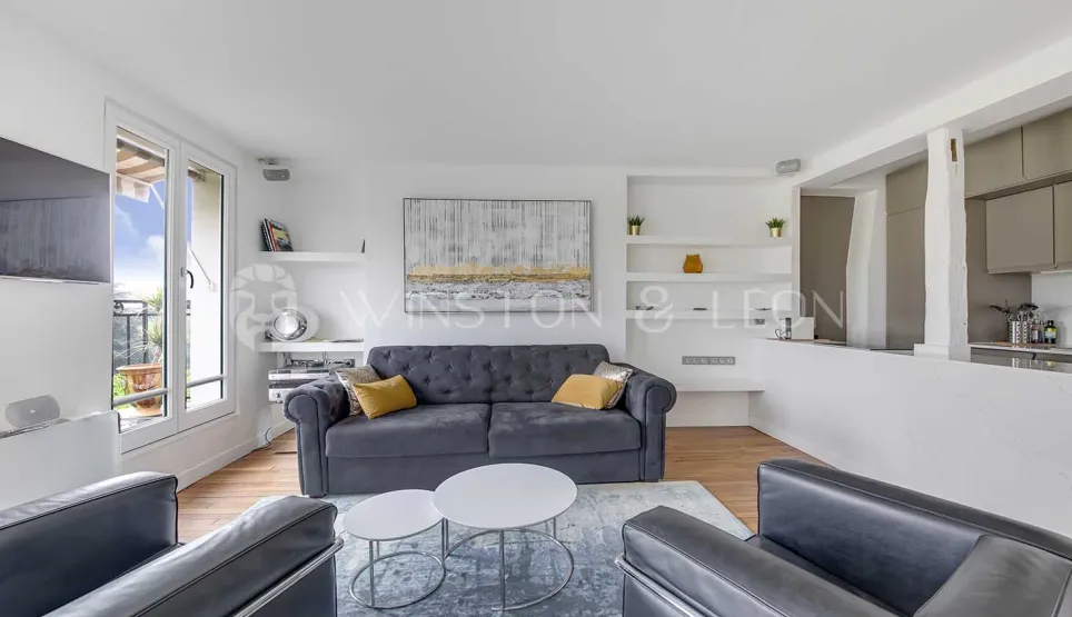 Salon moderne et lumineux d'un appartement de luxe avec canapé gris, balcon et cuisine ouverte. Design contemporain et épuré.