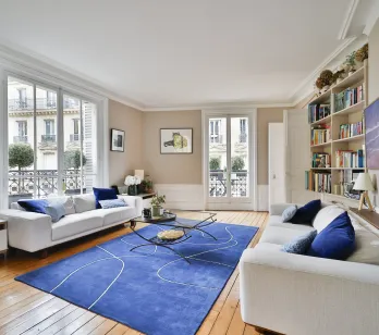 Salon de luxe parisien lumineux. Canapés blancs, tapis bleu moderne, parquet. Vue sur la ville. Élégance et confort haussmannien.
