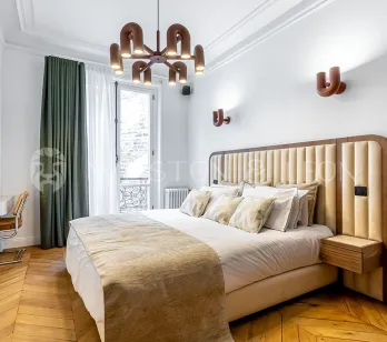 Chambre de luxe moderne avec tête de lit capitonnée, parquet chevrons, luminaires design et balcon. Élégance et confort pour un intérieur parisien.