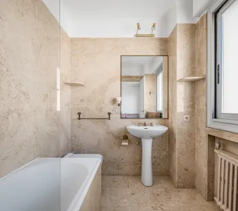 Salle de bain élégante en marbre beige, avec baignoire, lavabo sur colonne et miroir. Design classique et lumineux pour un intérieur de luxe.