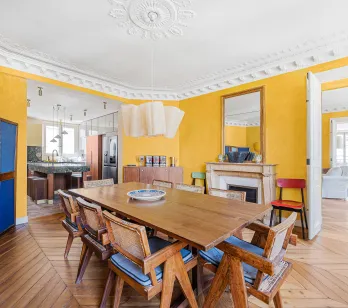 Élégante salle à manger parisienne aux murs jaunes, parquet chevron, cheminée. Appartement de luxe rénové, alliant charme ancien et design moderne.