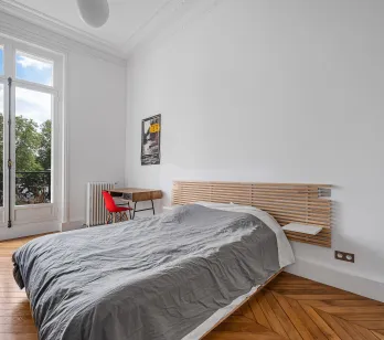 Chambre lumineuse avec balcon vue verdure, parquet chevrons et moulures. Design moderne et élégance parisienne pour un luxe discret.