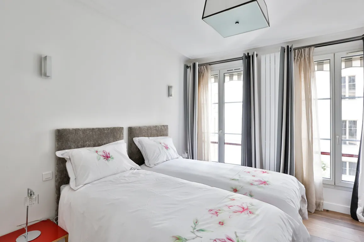 Chambre lumineuse et élégante avec deux lits jumeaux, draps fleuris, têtes de lit capitonnées. Grandes fenêtres et décoration moderne pour un confort de luxe.
