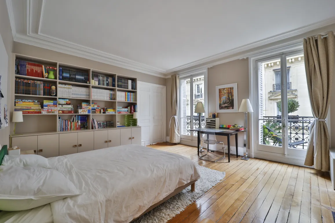 Chambre parisienne lumineuse avec parquet, moulures, bibliothèque intégrée et balcons. Élégance et confort pour un appartement de luxe.