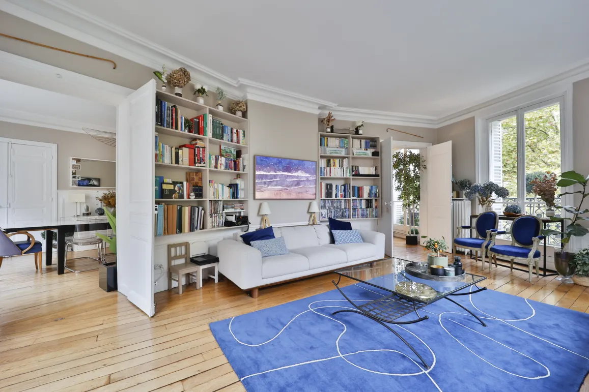 Salon de luxe parisien lumineux avec parquet, moulures, bibliothèque et vue verdure. Design élégant pour un appartement haussmannien spacieux.