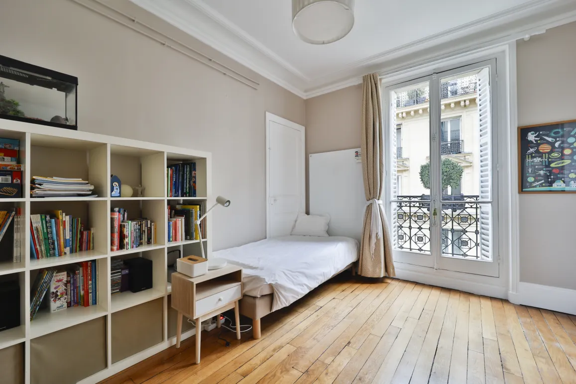Chambre lumineuse avec parquet, fenêtre française et balcon. Design épuré, espace optimisé. Idéal pour un studio parisien de luxe.