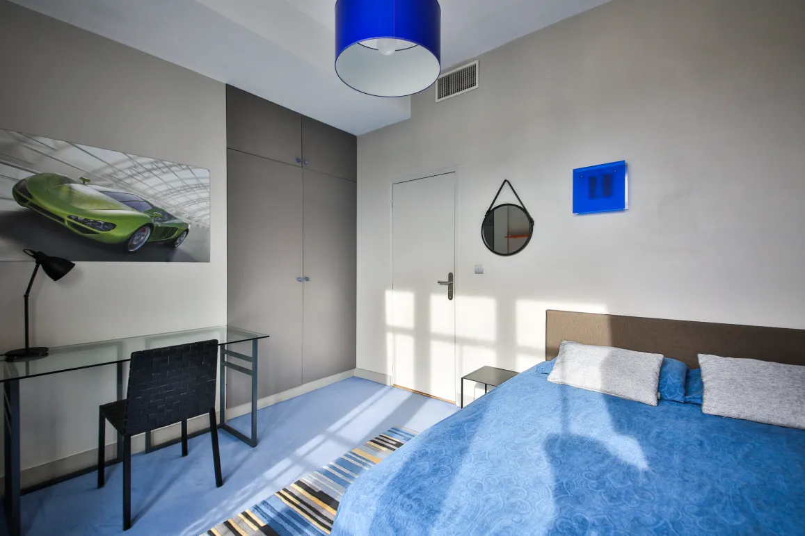 Chambre moderne et lumineuse, design épuré avec lit bleu, bureau verre, placards intégrés et œuvre d'art voiture sport. Ambiance chic.