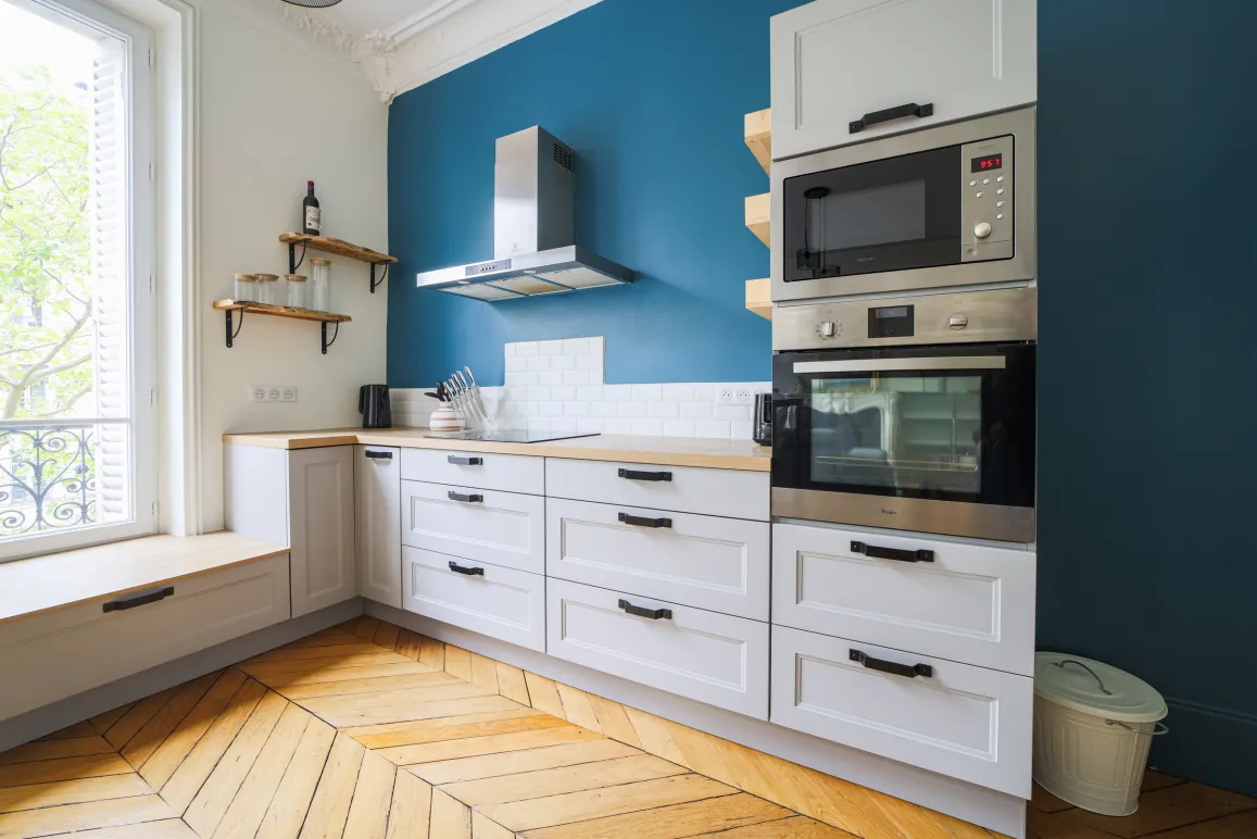 Cuisine moderne et lumineuse, murs bleus, parquet chevrons. Design élégant avec rangements blancs et électroménager intégré. Appartement parisien.