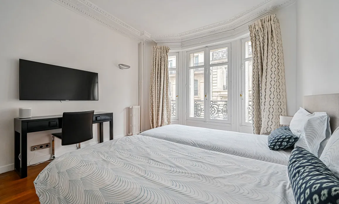 Chambre luxe parisienne, lits jumeaux, bureau, TV, grande fenêtre, moulures. Vue ville. Élégance et confort moderne.