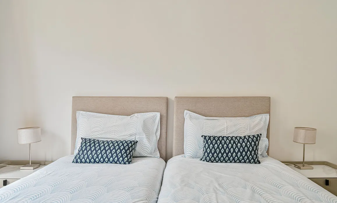 Chambre double luxe avec lits jumeaux, têtes de lit beige, literie épurée et coussins bleus. Design moderne et ambiance sereine pour un séjour raffiné.