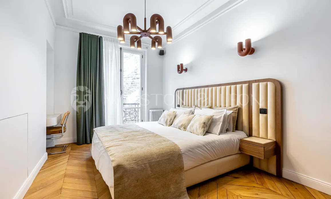 Chambre de luxe moderne avec tête de lit capitonnée, parquet chevrons, luminaires design et balcon. Élégance et confort pour un intérieur parisien.