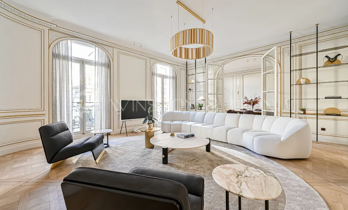 Salon Haussmannien parisien d'exception: moulures dorées, parquet point de Hongrie, canapé design blanc, mobilier luxe et lumière naturelle.