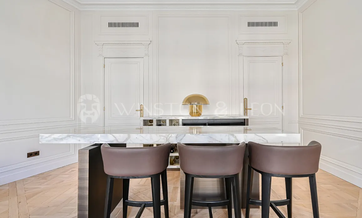 Cuisine de luxe avec îlot marbre, tabourets design. Boiseries élégantes, lampe dorée. Intérieur prestige, appartement parisien chic.