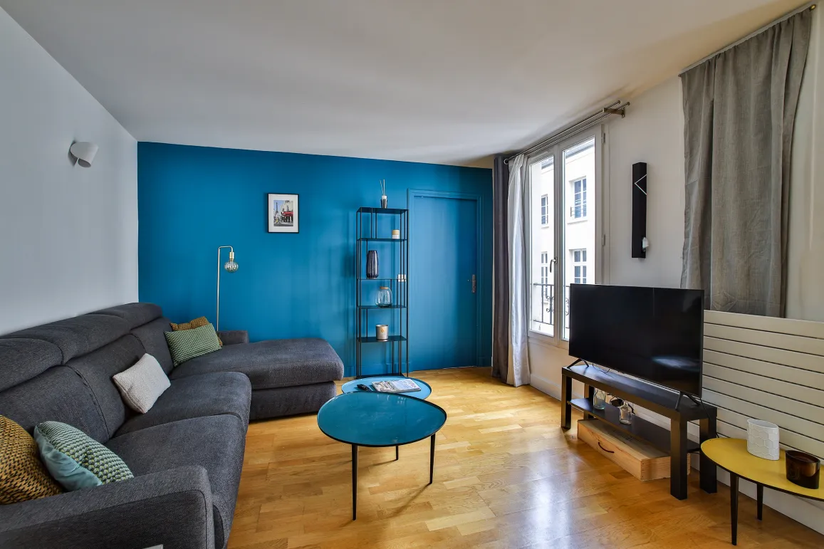 Salon moderne et lumineux avec mur bleu vibrant, canapé d'angle gris, parquet élégant. Idéal pour un appartement de luxe au design contemporain.