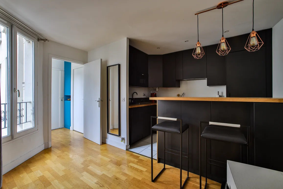 Cuisine moderne noire avec bar, tabourets design, parquet et luminaires cuivrés. Un espace de vie lumineux et élégant.