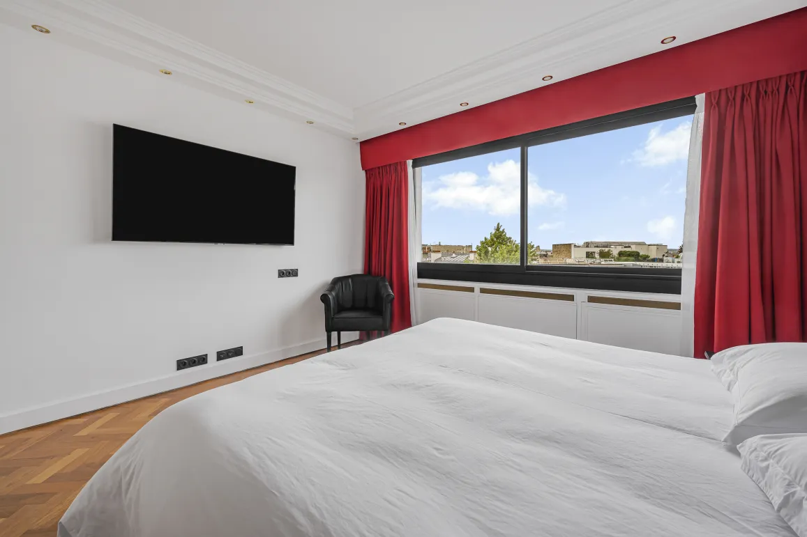 Suite parentale moderne et lumineuse avec vue citadine, TV, fauteuil et parquet. Le luxe et le confort d'un appartement haut de gamme.
