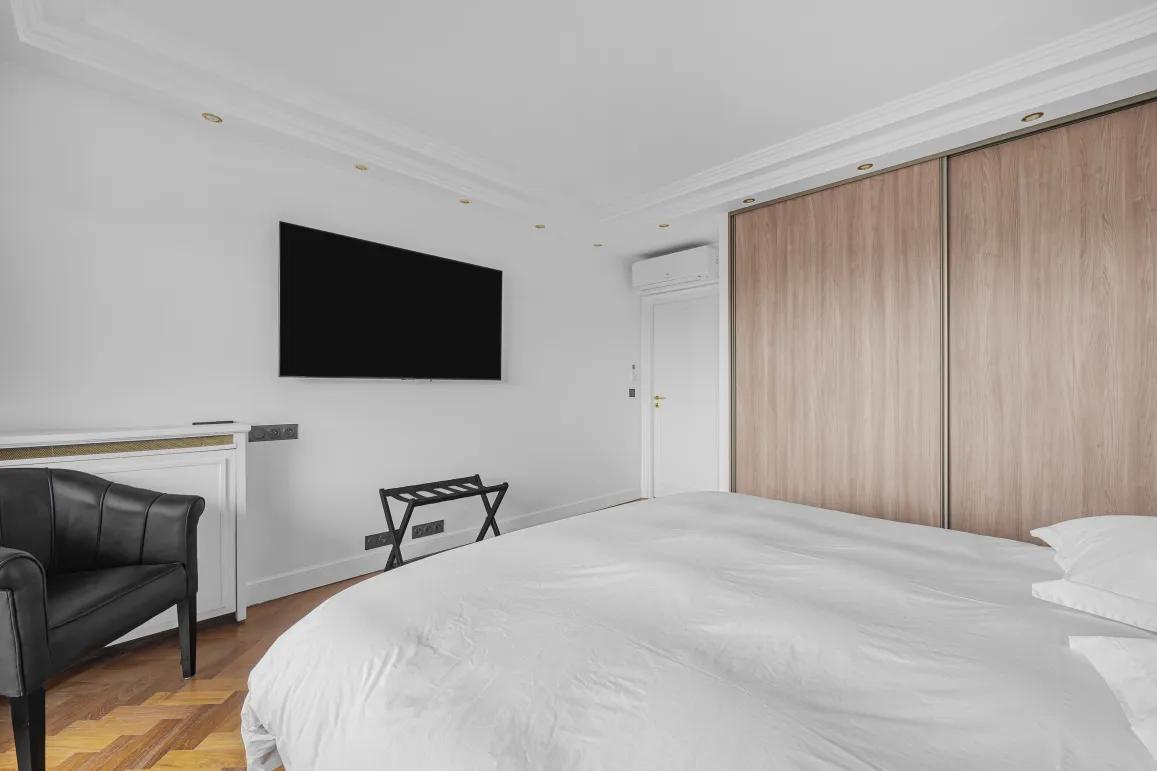 Chambre parentale moderne et élégante avec lit double, TV murale, dressing intégré bois, fauteuil design. Confort et luxe pour votre appartement.