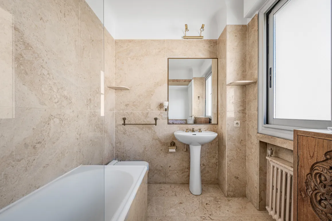 Salle de bain élégante en marbre beige, avec baignoire, lavabo sur colonne et miroir. Design classique et lumineux pour un intérieur de luxe.