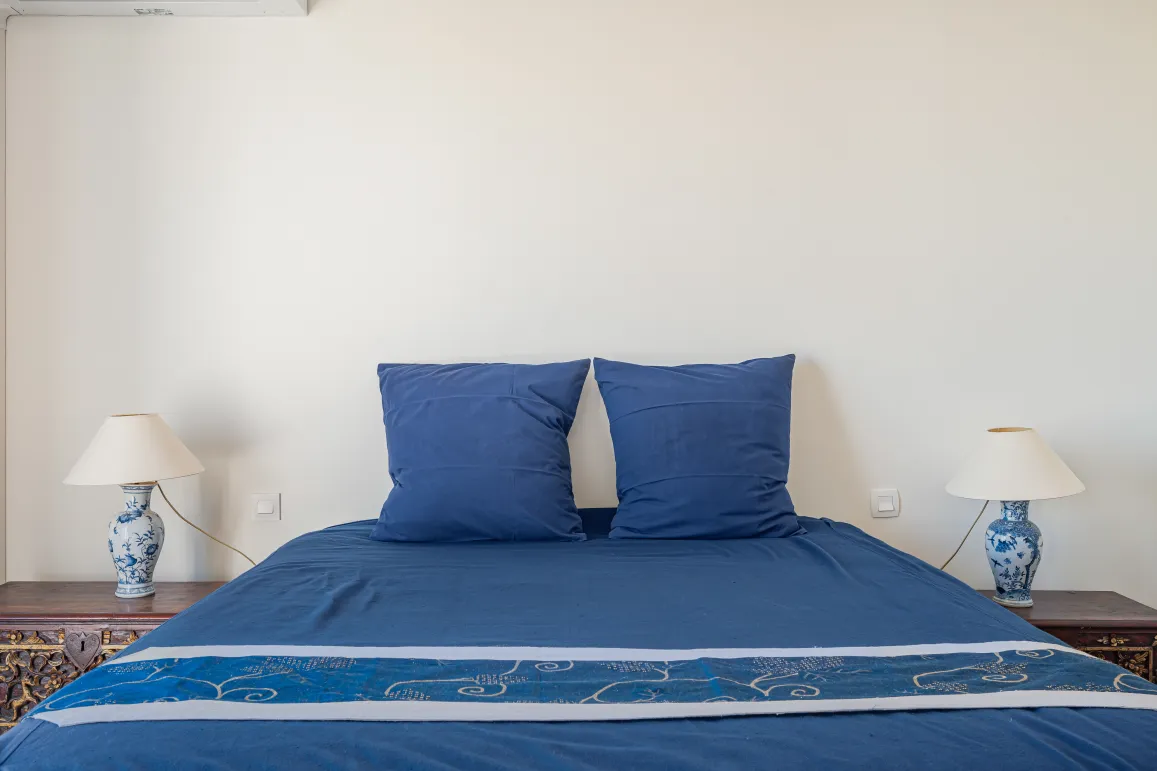 Chambre élégante et confortable avec lit bleu profond, tables de chevet sculptées et lampes en porcelaine. Ambiance raffinée pour propriété de luxe.
