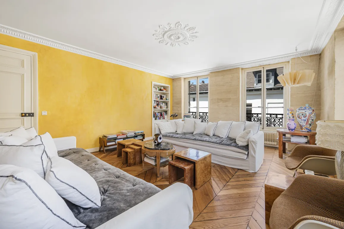Salon de luxe lumineux avec mur jaune texturé, parquet, moulures et canapés blancs. Élégance parisienne.