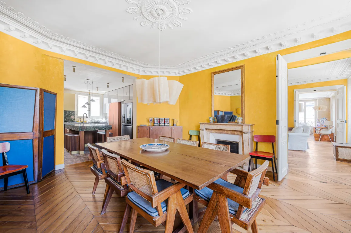 Élégante salle à manger parisienne aux murs jaunes, parquet chevron, cheminée. Appartement de luxe rénové, alliant charme ancien et design moderne.