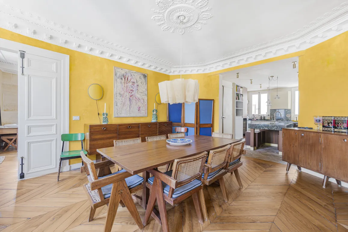 Appartement parisien de luxe: salle à manger lumineuse aux murs jaunes, parquet point de Hongrie, moulures. Cuisine ouverte design. Élégance vibrante.
