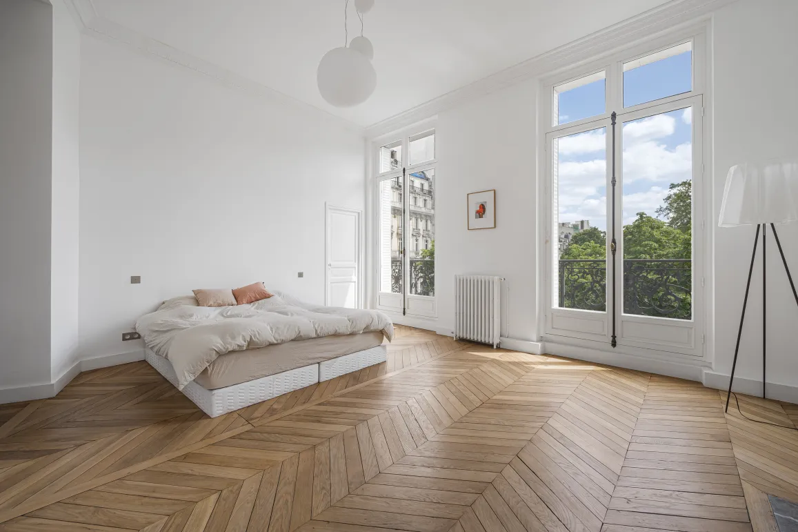 Chambre lumineuse d'appartement Haussmannien. Parquet chevrons, hauts plafonds, grandes fenêtres, vue verdure. Luxe parisien épuré.